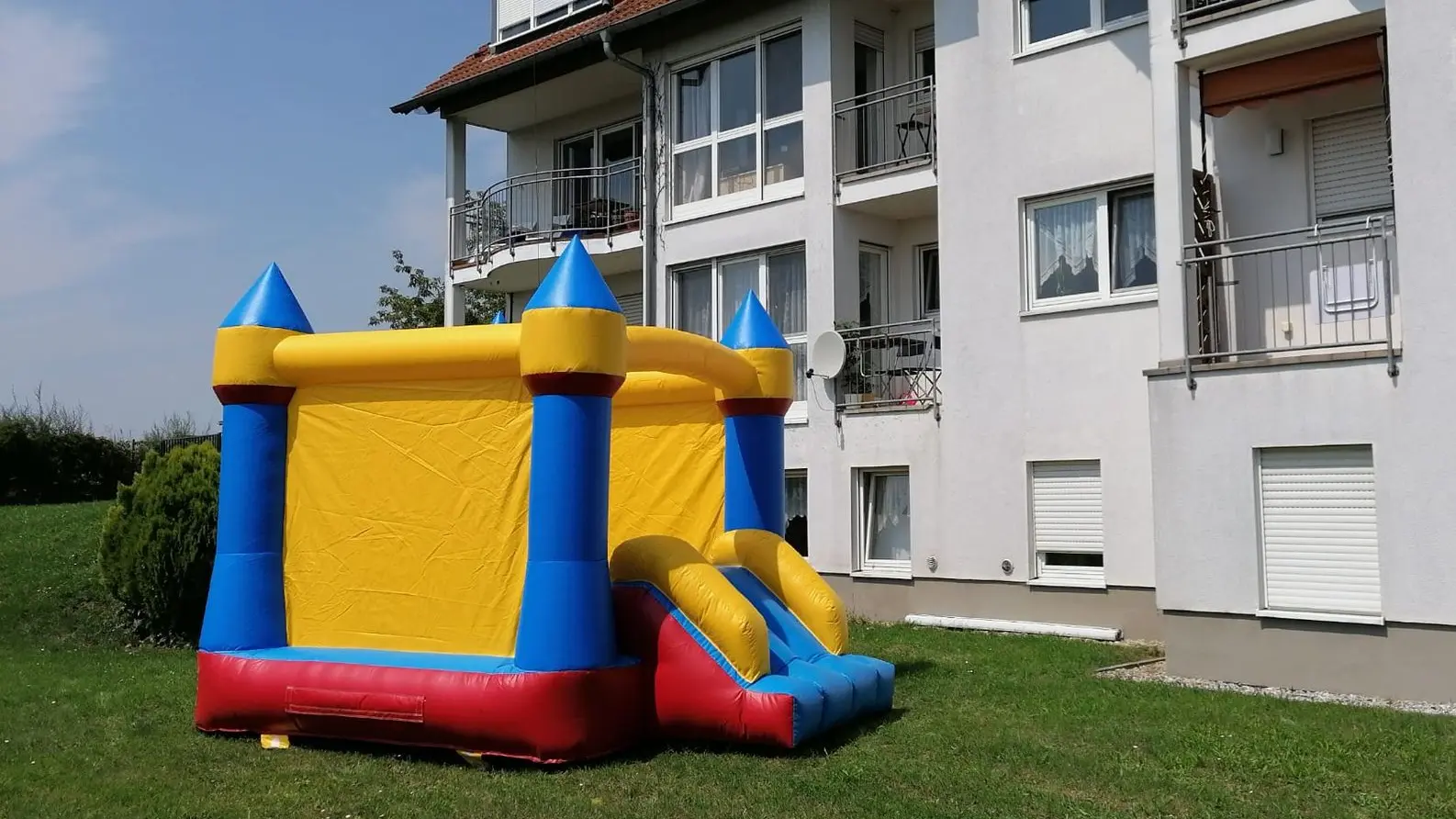 Abenteuerliche Hüpfburg mit Rutsche für Spiel im Freien! Bunte Hüpfburg mit Rutsche, auf einer Wiese neben einem Wohngebäude aufgestellt
