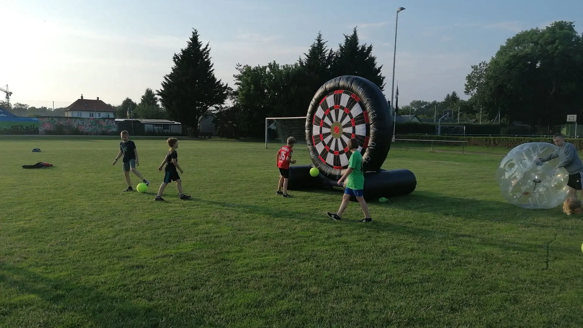 Aufregender Spaß im Freien mit aufblasbarer Dartscheibe und Spielaktivitäten! Kinder spielen mit einer großen aufblasbaren Dartscheibe, während andere mit Spielbällen auf dem Gras interagieren.